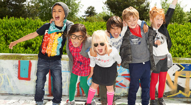 A group of six children, possibly at a Lille séminaire, stand outside, all smiling and cheering. They are dressed colorfully, with some wearing sunglasses, scarves, and a mix of casual clothing. Behind them are green bushes and trees. The ground is covered in colorful graffiti.
