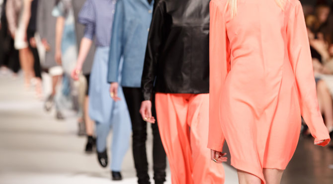 During a fashion show, a line of models walks down the runway wearing a variety of modern outfits, from a bright coral dress to black tops with orange pants and blue garments. The background is blurred, focusing on the models and their attire to increase collection visibility for PLM Empoli.