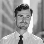 Black and white portrait of a man wearing a crisp white shirt and dark tie. He has neatly groomed dark hair and a short beard, and is facing the camera with a subtle smile. The background suggests an urban setting, hinting at faster market entry and speeding innovation in his field.
