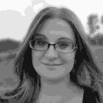 A black-and-white photograph of a woman with shoulder-length hair wearing glasses. She is smiling softly and looking directly at the camera. The background, slightly blurred, suggests an outdoor setting with trees and an open field, evoking a sense of serenity amidst speeding innovation.