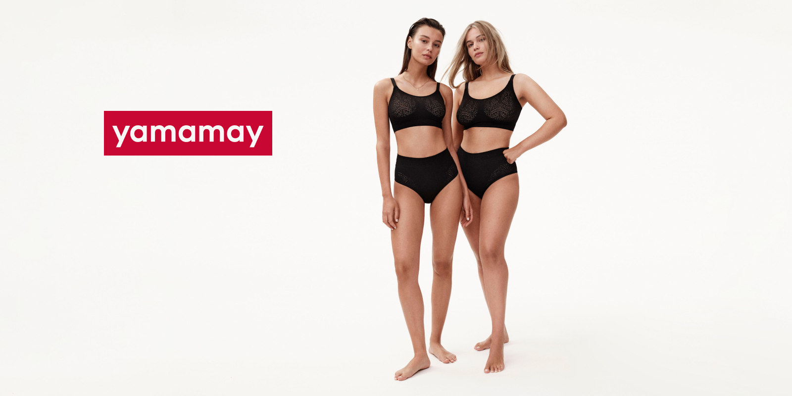 Two women standing side by side, modeling black lingerie sets against a plain white background. They are barefoot and positioned slightly angled towards each other. The word "Yamamay" appears in a red box to the left of the image.