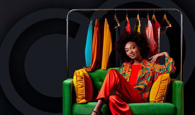 Fashionable woman sitting on a green sofa in front of a clothing rack showcasing vibrant, colorful garments.