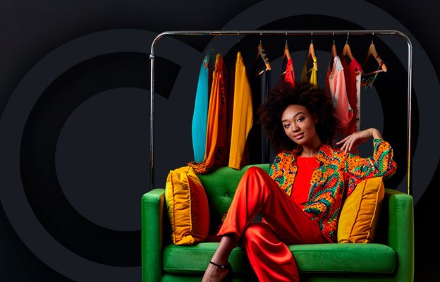Fashionable woman sitting on a green sofa in front of a clothing rack showcasing vibrant, colorful garments.