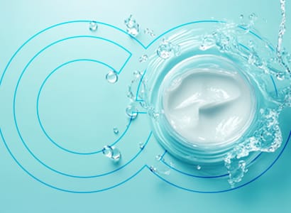 A jar of white cream is opened on a light blue background. Water splashes around and partially over the jar, creating a dynamic and fresh appearance. Circular blue lines are faintly overlaid on the background, mirroring how sustainability is integrated seamlessly into innovative product development for 2025.