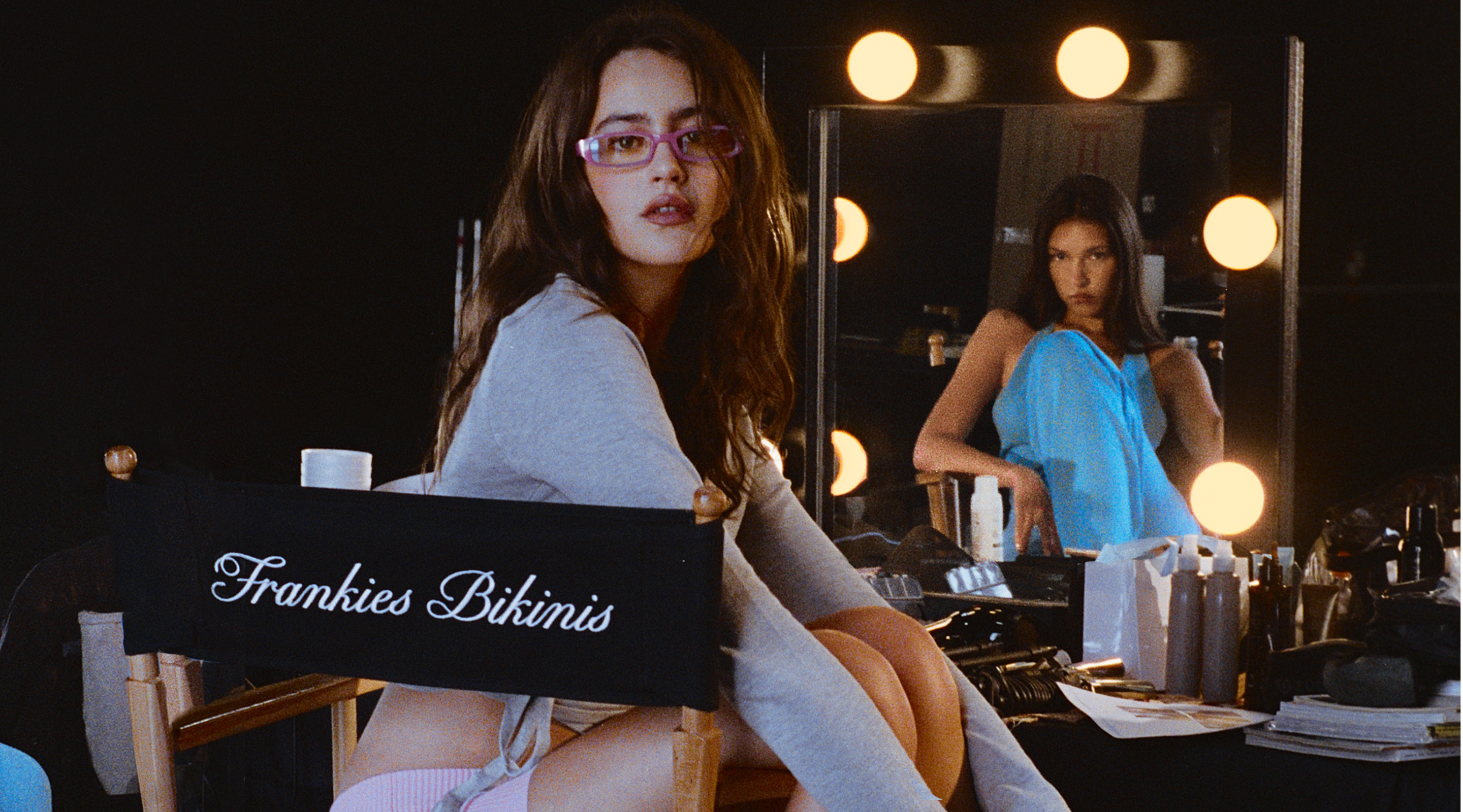 A woman in pink glasses sits on a chair labeled “Frankies Bikinis” in a makeup studio, showcasing product innovation, while another woman in blue is reflected in a mirror surrounded by bright lights and makeup products.