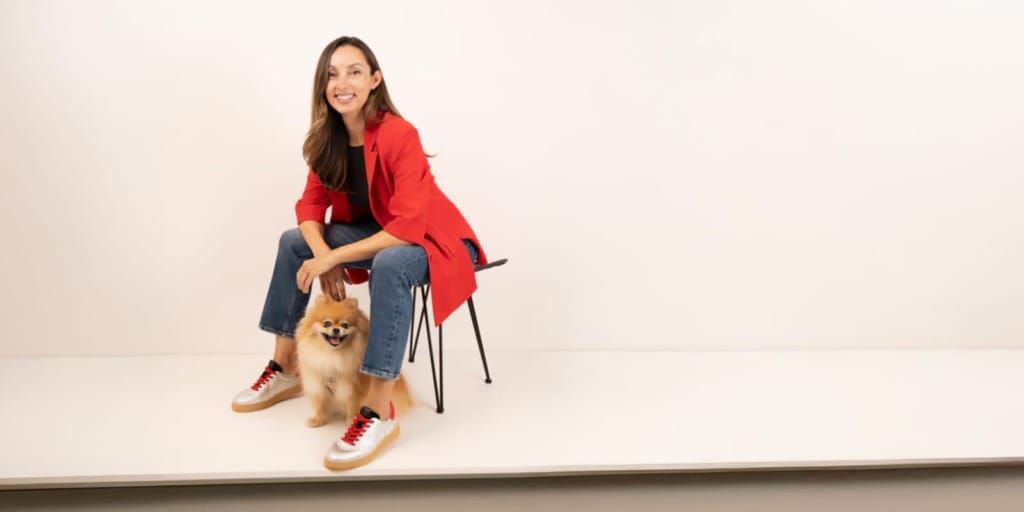 Kırmızı bir ceket ve kot pantolon giymiş gülümseyen bir kadın, düz beyaz bir arka plan önünde, ayaklarının dibinde mutlu bir şekilde oturan küçük tüylü bir köpekle birlikte bir taburede oturuyor.