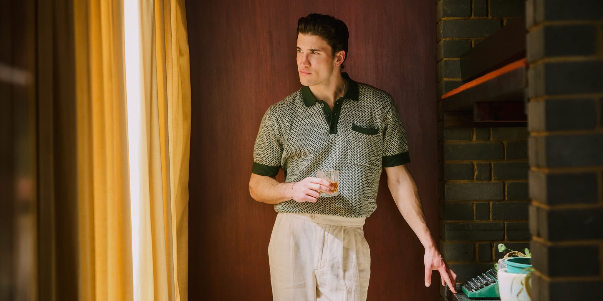 Một người đàn ông mặc áo sơ mi ngắn tay có hoa văn và quần âu màu sáng đứng bên cửa sổ có rèm màu vàng, cầm ly và nhìn ra ngoài, được bao phủ bởi ánh nắng ấm áp.