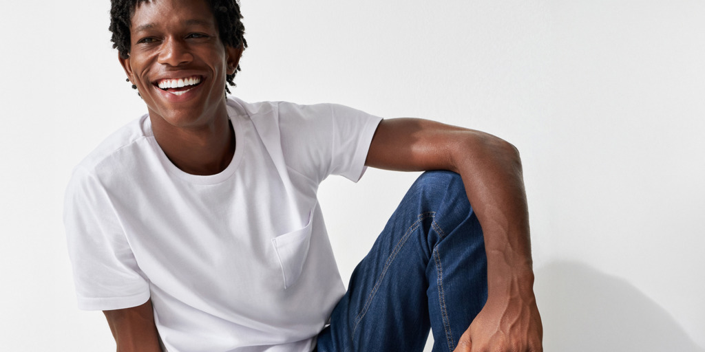 Uma pessoa sorridente, com cabelo curto e encaracolado, está sentada contra um fundo branco, vestindo uma T-shirt branca simples e calças de ganga azuis. Está a olhar para a câmara com uma expressão descontraída e alegre.